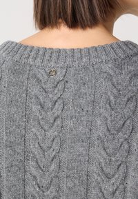 Pull en tricot gris avec un motif en relief, col à côtes et un petit logo métallique au dos. Apparence texturée et confortable.