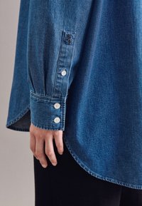Camicia di jeans a maniche lunghe, colore azzurro chiaro, con polsini abbottonati a tre bottoni bianchi e orlo curvo sul fondo.