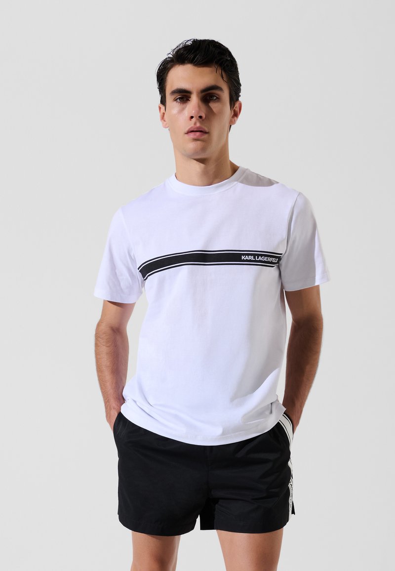 T-shirt bianco in cotone con una striscia orizzontale nera che presenta il testo "KARL LAGERFELD". Maniche corte, collo a giro e fit casual.