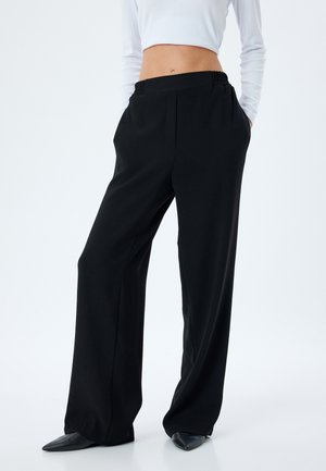 Zwarte wijde broek met een gladde textuur, elastische tailleband en zijzakken, gecombineerd met glanzende zwarte puntschoenen.