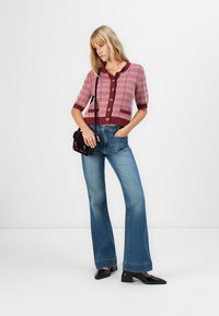 Femme portant un cardigan à manches courtes boutonné à carreaux roses, un pantalon en jean bleu à jambes larges, des chaussures noires, et portant un petit sac à épaule noir.