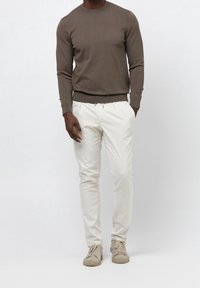 Man med brun långärmad tröja, off-white byxor och beige sneakers som står med händerna i fickorna.