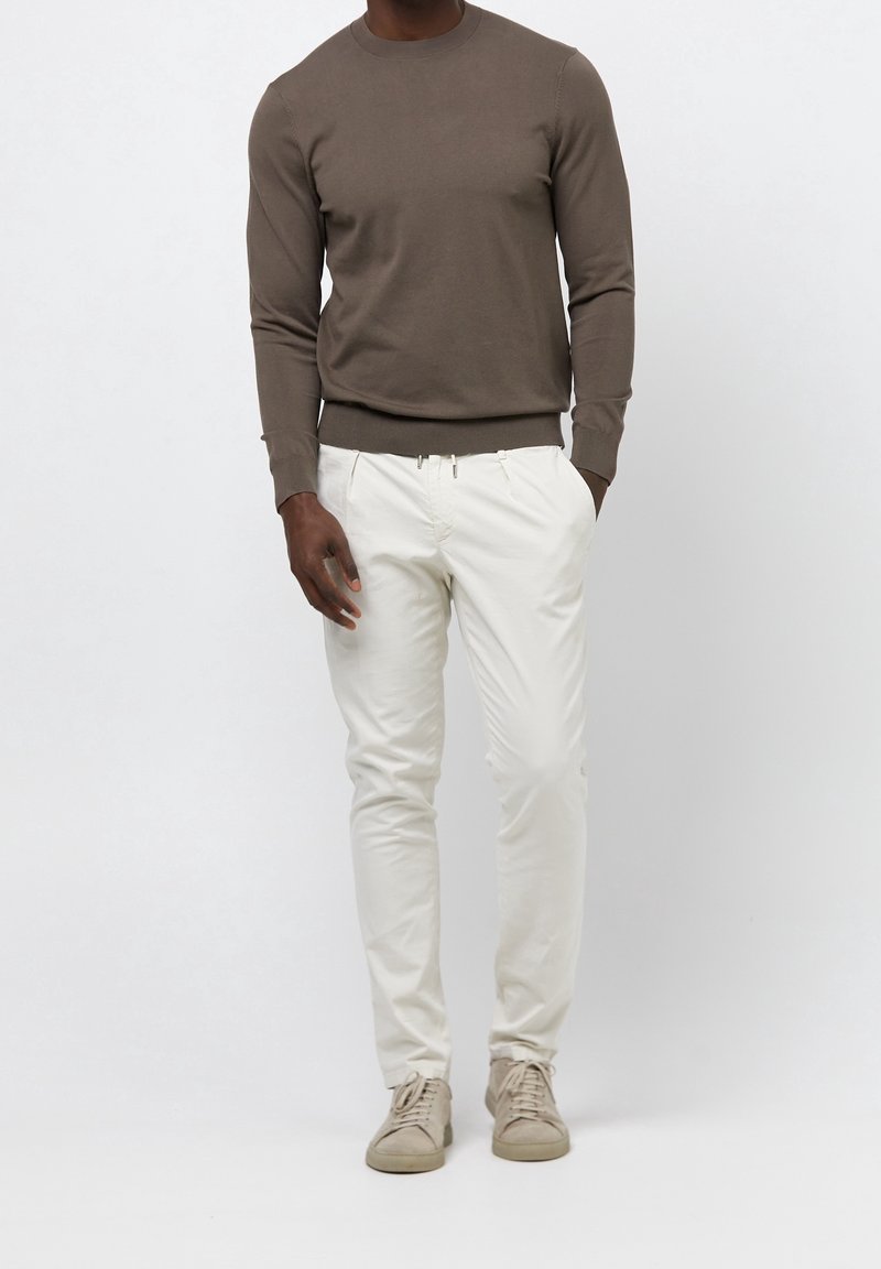 Man med brun långärmad tröja, off-white byxor och beige sneakers som står med händerna i fickorna.