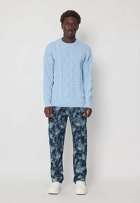 Pull en tricot bleu clair, pantalon abstrait bleu foncé à motifs et baskets blanches. L'ensemble présente une coupe décontractée et des matériaux texturés.