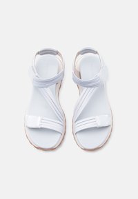 Paire de sandales blanches avec des brides croisées élastiques et des Fermetures Velcro, semelles plates, vues de dessus sur un fond clair.