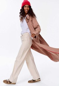 Kobieta idąca, ubrana w czerwoną czapkę beanie, długi beżowy płaszcz z czerwonymi wykończeniami, białą koszulę, kremowe spodnie i trampki w cętki.