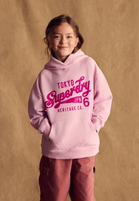 Felpa con cappuccio rosa con tasca a marsupio, decorata con testo bianco e rosa "Tokyo Superdry JPN 6 Heritage Co." Abbinata a pantaloni cargo color rosa.