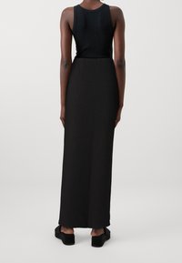 Schwarzes ärmelloses Oberteil und lange schwarze Rockkombination. Der Stoff wirkt glatt mit einer leichten Textur. Tragen mit einfachen schwarzen Plateauschuhen.