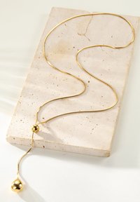 Goldene Halskette mit einem glatten, runden Perlenakzent und einem spitz zulaufenden Tropfenvorhang, die auf einer strukturierten, beigen Kalksteinoberfläche ruht.