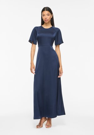 Femme portant une robe longue en satin bleu marine avec des manches courtes flottantes, debout droite devant un fond blanc.