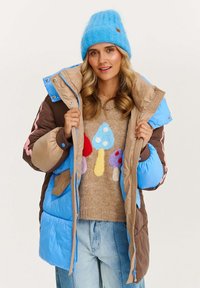Žena s modrou čiapkou, modro-hnedou zateplenou bundou, béžovým sveterom so farebným dizajnom húb a svetlomodrými džínsami.