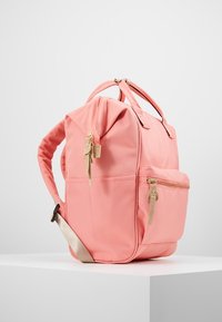 Růžový látkový batoh s pevnou strukturou, vybavený horním madlem, nastavitelnými ramenními popruhy a přední kapsou na zip. Zlaté kovové doplňky.