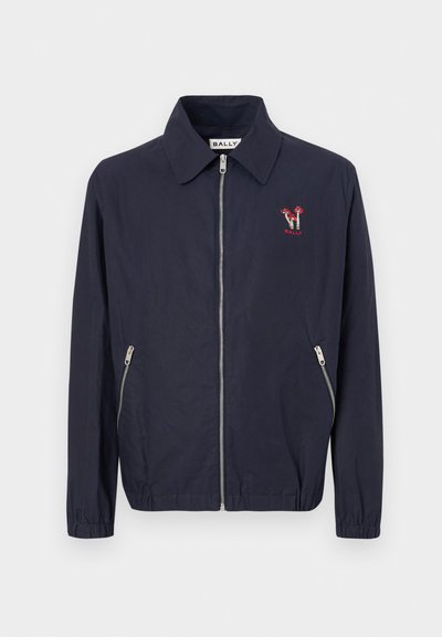 Tumed sinine kerge jakker, millel on pikad varrukad, lukuga kinnitamine, küljetaskud ja punane tikand logoga vasakul rinnal.