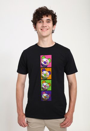Jonge man draagt een zwart T-shirt met vier kleurrijke clownsgezichten die verticaal gestapeld zijn aan de voorkant tegen een witte achtergrond.