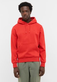 Mężczyzna w jasnoczerwonej bluzie z kapturem, z przednią kieszenią i zielonych spodniach, stojący na tle jednolitego jasnego tła.