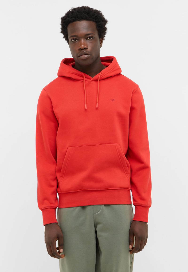 Mężczyzna w jasnoczerwonej bluzie z kapturem, z przednią kieszenią i zielonych spodniach, stojący na tle jednolitego jasnego tła.