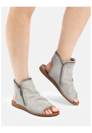 Beine in grauen, offenen Ledersandalen mit seitlichen Reißverschlüssen und flachen braunen Sohlen, Fersen und Zehen sind vor weißem Hintergrund sichtbar.