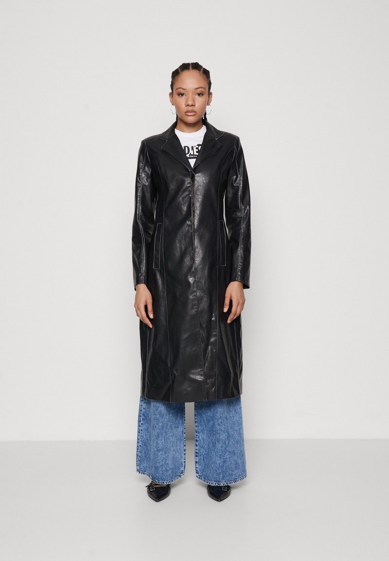Abrigo de cuero negro con solapa de muesca, cierre de botón, bolsillos laterales y costuras blancas en contraste, combinado con jeans azules de pierna ancha.