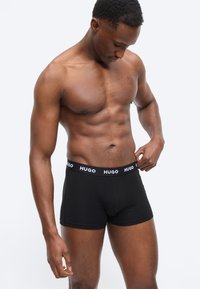 Μυώδης άνδρας που διορθώνει μαύρα μπόξερ με λογότυπο "HUGO" στη μέση, στέκεται μπροστά σε απλό ανοιχτόχρωμο φόντο.