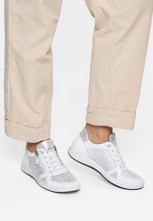 Person trägt beige Cordhosen und weiße Turnschuhe mit silbernen Akzenten und steht vor einem weißen Hintergrund.