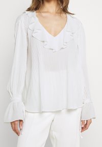 Camicia bianca con colletto arricciato e texture plissettata; maniche lunghe con polsini svasati. Tessuto morbido e leggero. Design semplice ed elegante.