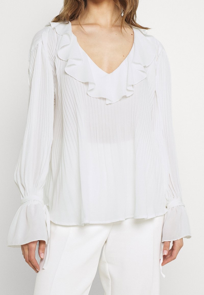Camicia bianca con colletto arricciato e texture plissettata; maniche lunghe con polsini svasati. Tessuto morbido e leggero. Design semplice ed elegante.