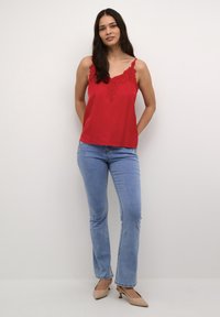 Rote Satin-Camisole mit Spitzenrand und dünnen Trägern. Kombiniert mit hellblauen Schlagjeans und beigen High-Heels. Glatte Textur und einfaches Design.