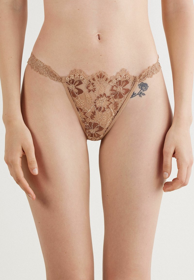 String en dentelle de couleur beige avec des motifs floraux. Présente des bords festonnés et une délicate bordure en dentelle le long de la taille. Texture douce.