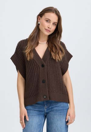 Femme aux longs cheveux bruns porte un cardigan sans manches en grosse maille marron foncé et un jean bleu, sur un fond clair uni.