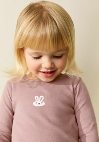 Fată tânără blondă care privește în jos zâmbind, purtând un tricou roz cu dungi, cu o mică broderie albă de iepuraș pe piept.