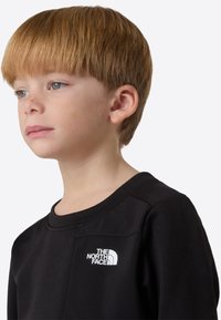 Musta huppari, jossa The North Face -logo valkoisena rinnassa, pyöreä kaula-aukko ja pehmeä pinta, jota lapsi käyttää.