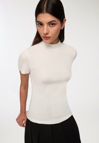 Biała, ribbrowana bluzka z krótkim rękawem o wysokim dekolcie, dopasowany krój i gładka faktura. W zestawie z czarną plisowaną spódnicą.
