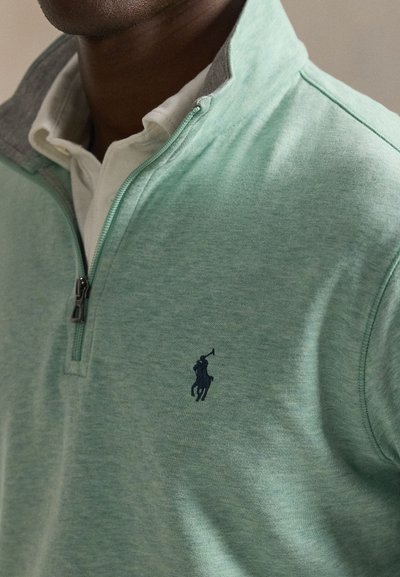 Sweatshirt verde com fecho de correr, gola cinzenta clara, apresenta um logótipo de pólo azul-marinho no peito e um tecido macio e texturizado.