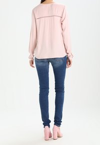 Ljusrosa långärmad topp med rund halsringning och dekorativ sömnad på baksidan, i kombination med blå skinny jeans och rosa klackar.