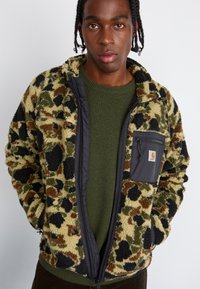 Veste léopard floue, de couleur vert olive et marron, avec des accents noirs, des poches zippées et une texture douce, comportant un zip avant et un col haut.