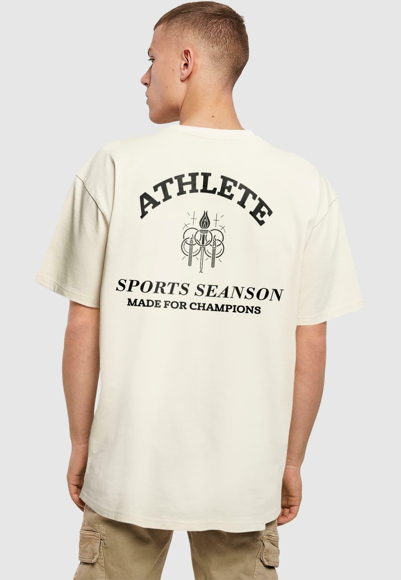 Kremkrāsas pārāk liels t-krekls ar melnu uzrakstu: "SPORTISTS" un grafiku zem tā, kas norāda: "SPORTA SEZONA VEIDOTA ČEMPIONIEM." Īsas piedurknes.