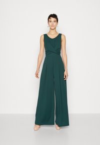 Une combinaison verte en tissu doux avec un décolleté en V, un détail torsadé à la taille, des jambes larges, et associée à des chaussures beige clair.