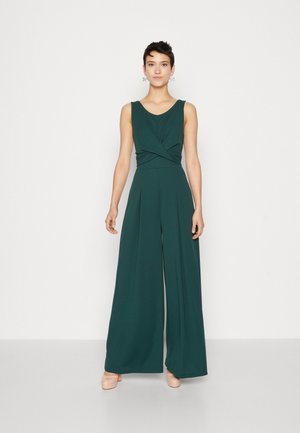 Une combinaison verte en tissu doux avec un décolleté en V, un détail torsadé à la taille, des jambes larges, et associée à des chaussures beige clair.