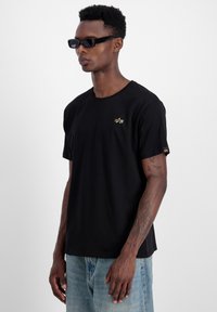 Schwarzes T-Shirt aus Baumwolle mit einem einfachen Rundhalsausschnitt, kurzen Ärmeln und einem goldenen Logo auf der Brust. Kombiniert mit hellblauen Jeans.