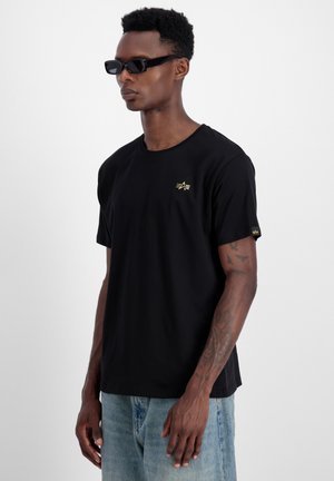 Schwarzes T-Shirt aus Baumwolle mit einem einfachen Rundhalsausschnitt, kurzen Ärmeln und einem goldenen Logo auf der Brust. Kombiniert mit hellblauen Jeans.