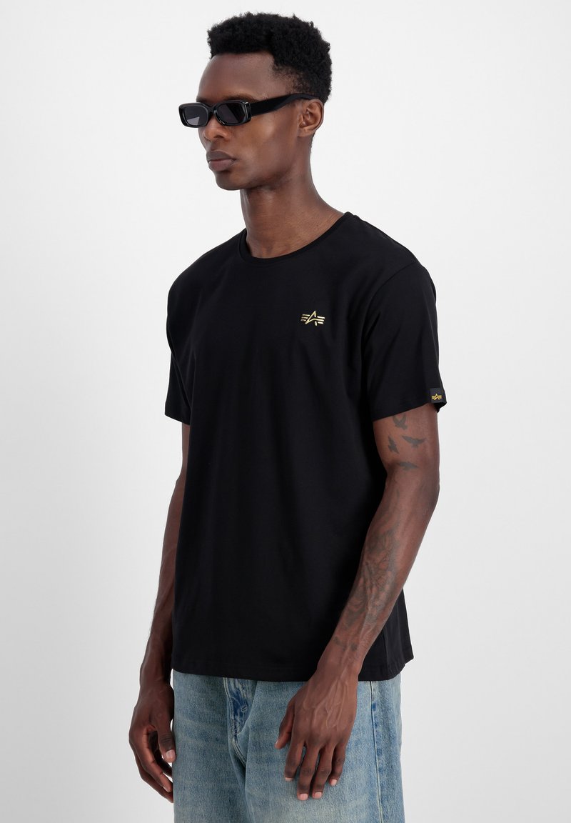 Schwarzes T-Shirt aus Baumwolle mit einem einfachen Rundhalsausschnitt, kurzen Ärmeln und einem goldenen Logo auf der Brust. Kombiniert mit hellblauen Jeans.
