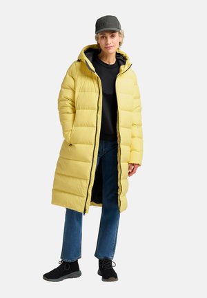 Geel gewatteerde lange jas met capuchon, voorzien van een rits, zwarte accenten en zijzakken. Gecombineerd met donkere jeans en zwarte schoenen.