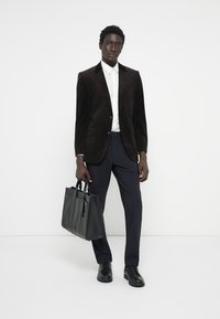 Dunkelbrauner Kordblazer über einem weißen Hemd mit Knöpfen, kombiniert mit marineblauen Hosen und glänzenden schwarzen Schuhen, hält eine schwarze Leder-Shopper. 