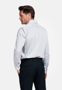 Chemise blanche à motif avec un col écarté, des poignets à boutons et de petits motifs géométriques en bleu et rouge. Associée à un pantalon noir.