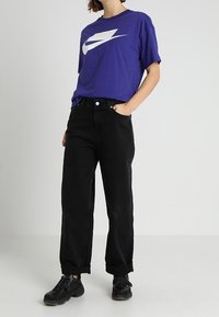 Osoba ubrana w fioletową koszulkę Nike, czarne spodnie z wysokim stanem i mankietami oraz czarne tenisówki, stojąca z jedną ręką w kieszeni na jednolitym tle.