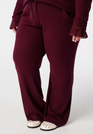 Personne portant un pantalon en tricot côtelé bordeaux et un haut assorti avec des poignets en dentelle, se tenant sur un fond clair avec des chaussures blanches à clous.