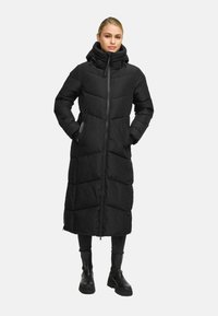 Femme portant un manteau d'hiver long noir rembourré avec un col haut, une fermeture éclair sur le devant, les mains dans les poches, un pantalon noir et des bottes noires.