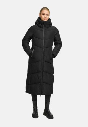 Femme portant un manteau d'hiver long noir rembourré avec un col haut, une fermeture éclair sur le devant, les mains dans les poches, un pantalon noir et des bottes noires.