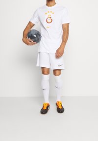 Uomo in divisa da calcio bianca che tiene in mano un pallone nero, indossando scarpe da calcio nere e arancioni, calze bianche e una maglietta con il logo di una squadra sportiva e stelle.