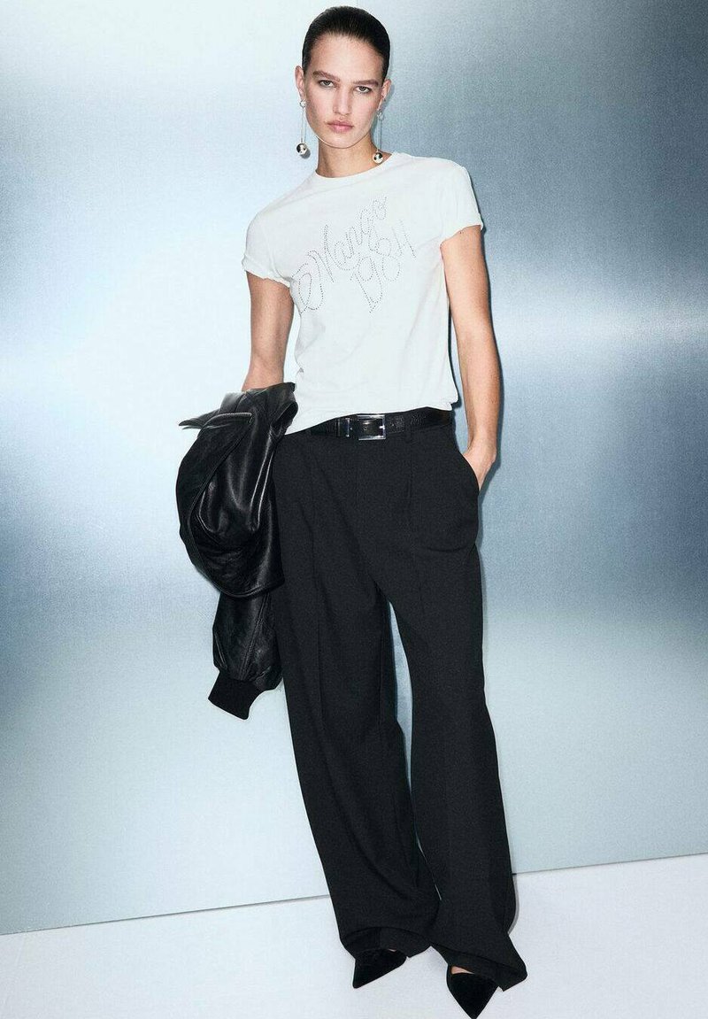 Witte T-shirt met bedrukking, zwarte high-waisted broek, zwarte leren jas en puntige zwarte hakken. Zilveren oorhangers.